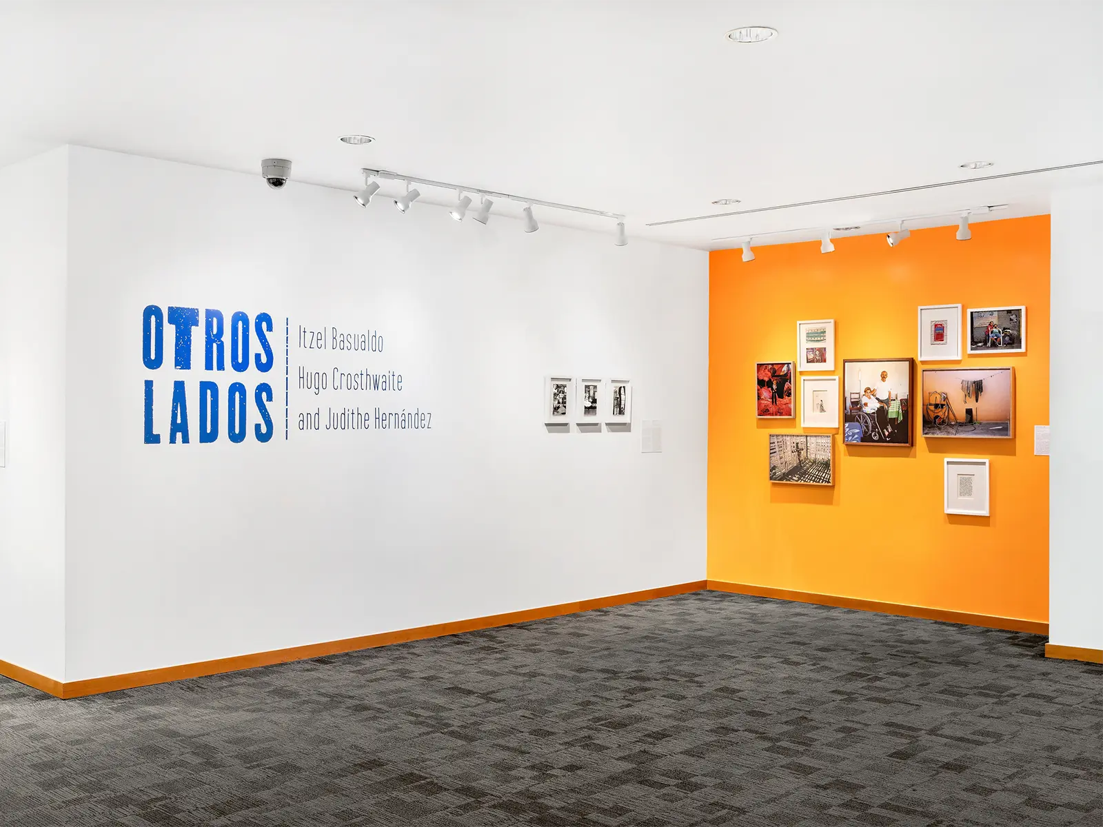 Art exhibition at Frost Art Museum featuring framed artworks on white and orange walls with text ‘OTROS LADOS’ and artist names